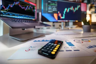 smartphone calculator on desk with financial charts behind by Jakub Żerdzicki courtesy of Unsplash.
