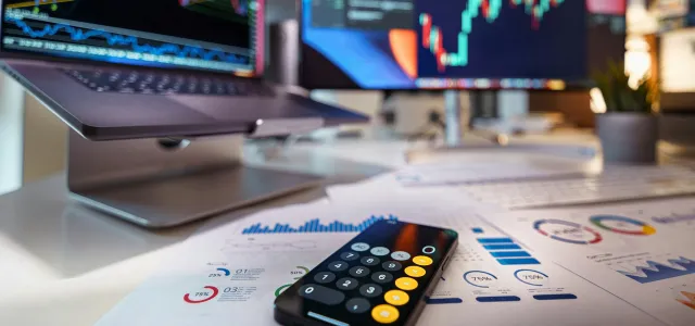 smartphone calculator on desk with financial charts behind by Jakub Żerdzicki courtesy of Unsplash.